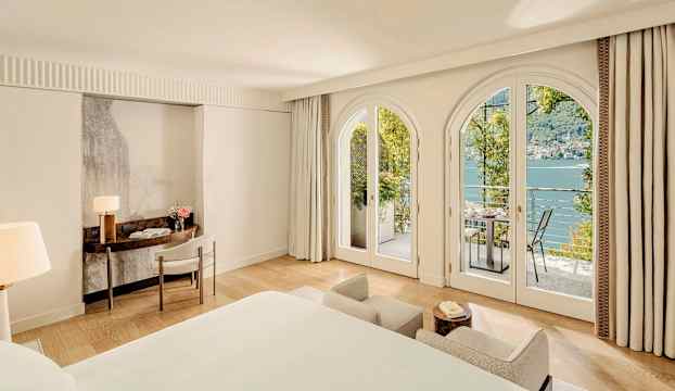 Lyxigt hotellrum med stora välvda fönster som öppnar upp mot en balkong med utsikt över en sjö. (AI-genererad text)