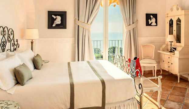 Ett elegant sovrum med dubbelsäng, två nattduksbord, väggdekorationer och ett skrivbord med spegel, fönster med havsutsikt. (AI-genererad text)