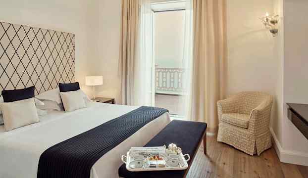 Ett sovrum med en stor säng, mörka och ljusa textilier, en sittbänk, en stol och ett litet soffbord bredvid balkongdörrar med vita gardiner. (AI-genererad text)