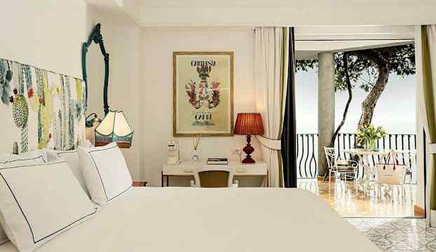 Ett elegant sovrum med en säng, skrivbord, bekväma möbler och en balkong som vetter mot havsutsikt. (AI-genererad text)