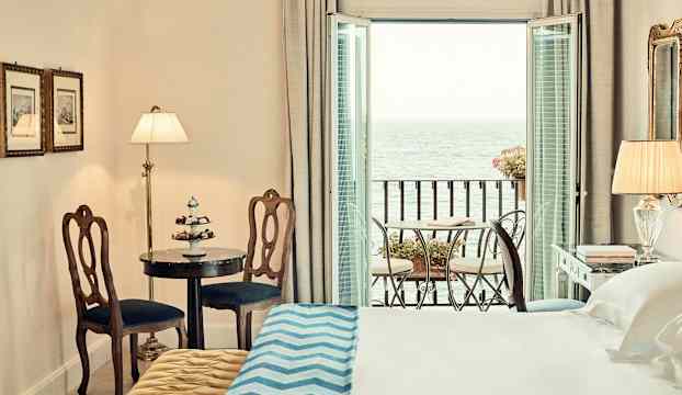 Ett hotellrum med en säng, bord med stolar och en balkong med utsikt över havet. (AI-genererad text)