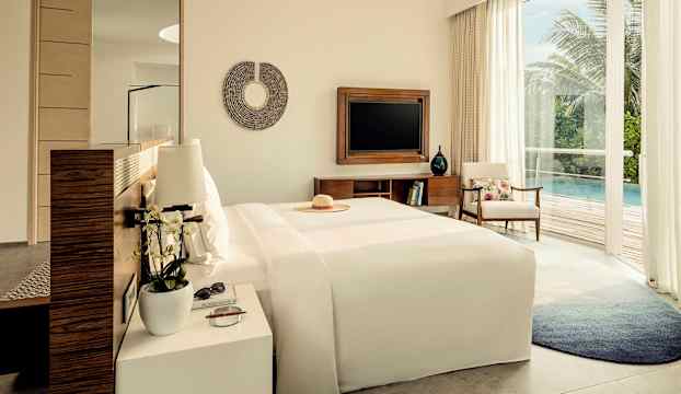 Ett hotellrum med stor säng, sidobord och TV. Utsikt mot en terrass med pool och grönska. (AI-genererad text)