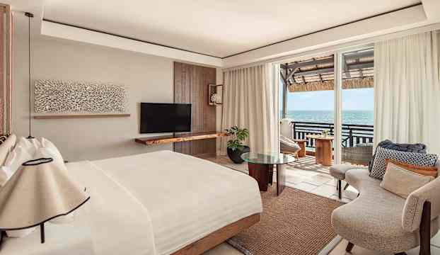 Ett elegant hotellrum med en stor säng, en fåtölj och utsikt över havet från en balkong. (AI-genererad text)