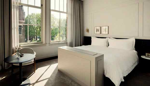 Ett ljust och elegant hotellrum med en stor säng, stora fönster och ett litet runt bord. (AI-genererad text)