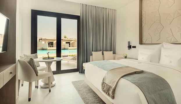 Ett elegant hotellrum med en dubbelsäng, sittgrupp, bord och stolar, med utsikt över en pool genom glasdörrar. (AI-genererad text)