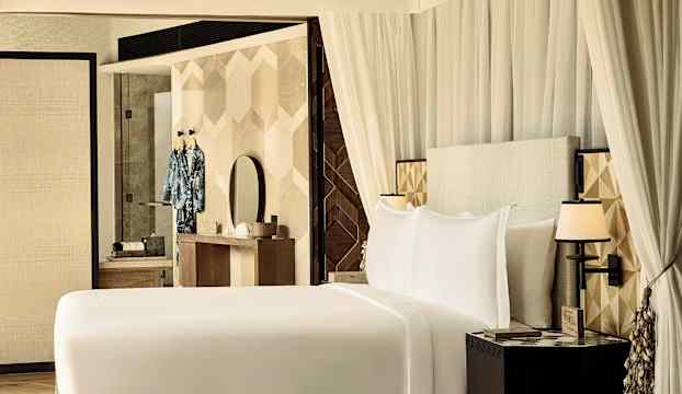 Ett hotellrum med en stor säng, vita sängkläder och en dekorerad sänggavel, samt ett skrivbord med spegel och kläder hängande på väggen. (AI-genererad text)