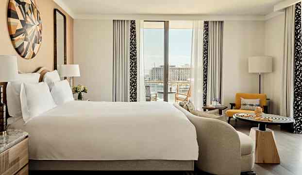 Ett hotellrum med stor säng, sittmöbler och en utsikt mot en marina genom stora fönster. (AI-genererad text)