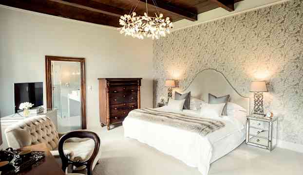 Ett elegant sovrum med en dubbel säng, dekorativa lampor och en detaljerad tapet på väggen. (AI-genererad text)