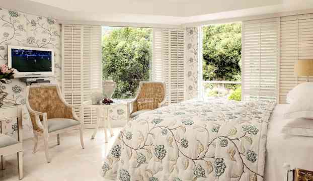 Ett rum med en vit säng, blommigt överkast, två stolar och ett litet bord. Fönster med utsikt mot grönska. (AI-genererad text)