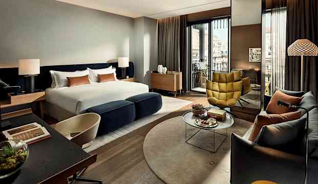 Ett modernt hotellrum med stor säng, sittgrupp och utsikt mot byggnader. (AI-genererad text)