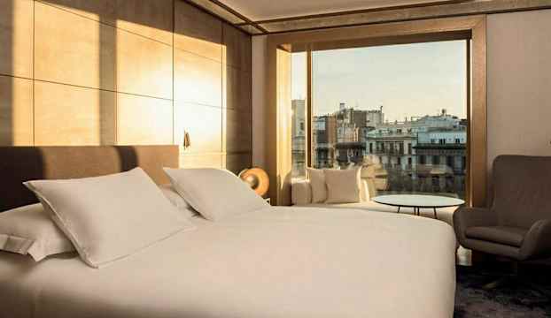 Hotellrum med en stor säng, fåtölj och utsikt över en stad. (AI-genererad text)