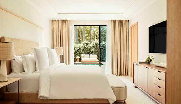 Ett modernt hotellrum med en dubbelsäng, en TV på väggen och ett stort fönster med utsikt mot en möblerad terrass och grönska. (AI-genererad text)