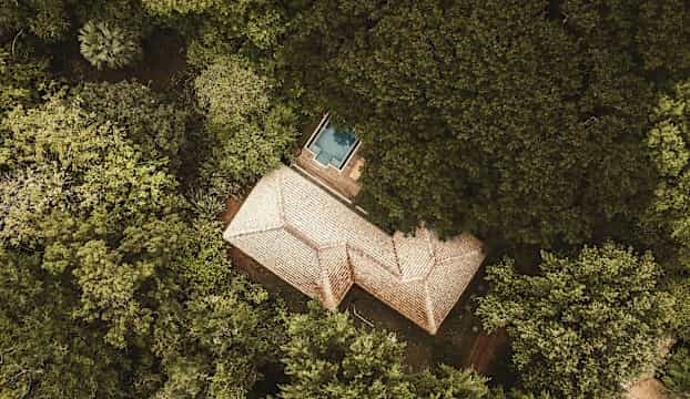 Luftperspektiv av en byggnad med tegeltak omgiven av tät grönska, en liten blå pool syns nära byggnaden. (AI-genererad text)