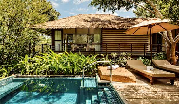 Avbildning av en liten pool framför en bungalow med halmtak, omgiven av grönska. (AI-genererad text)
