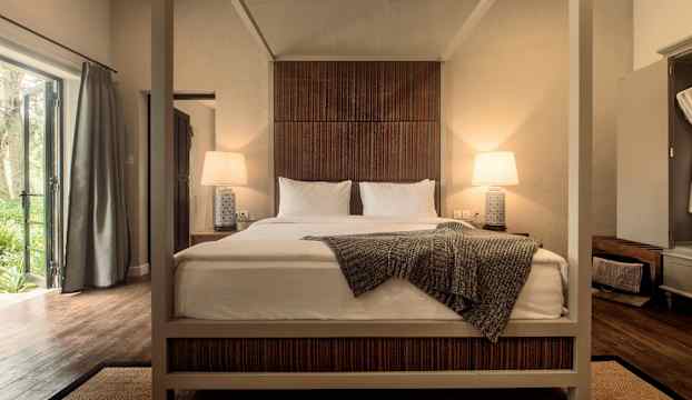 En säng med pelarram står centralt i ett rum, omgivet av öppna fönster och belysning vid sängborden. (AI-genererad text)