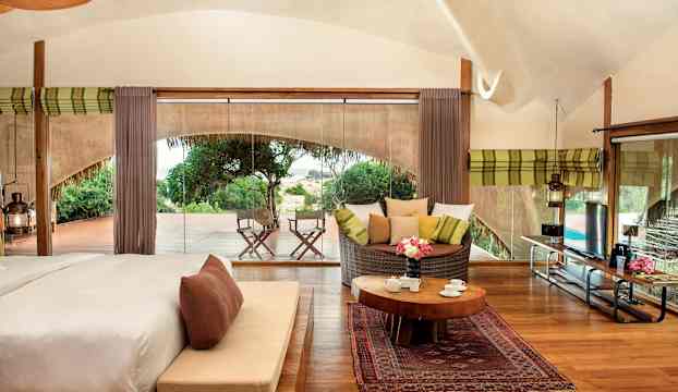 Ett rum med säng, sittgrupp, träbord och utsikt via glasvägg mot en terrass och grönska. (AI-genererad text)