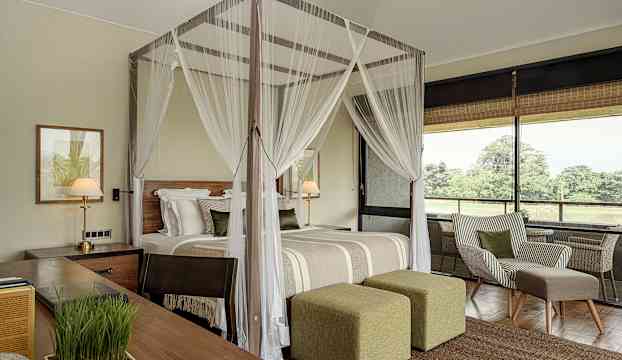 Ett hotellrum med himmelssäng, stol och utsikt genom fönster mot grönska. (AI-genererad text)