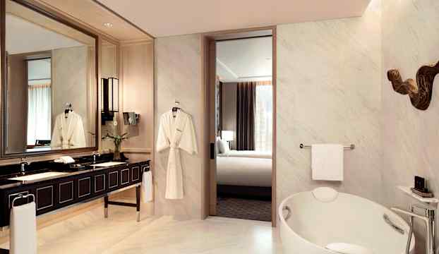 Lyxigt badrum med marmorgolv, ovalt badkar, dubbelhandfat med mörk bänkskiva och spegel samt utsikt mot ett sovrum. (AI-genererad text)