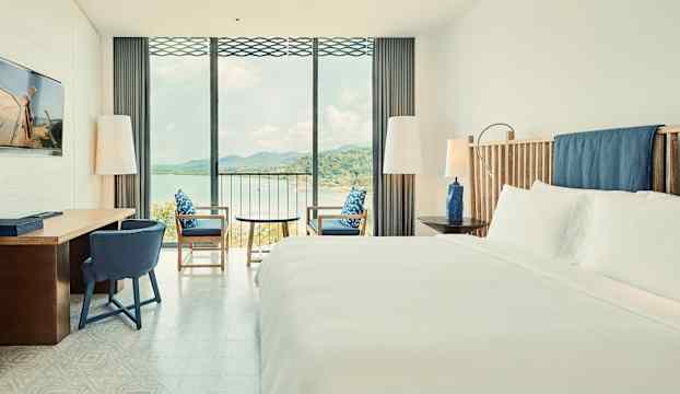 Ett hotellrum med en stor säng och sittgrupp som har utsikt mot havet genom stora fönster. (AI-genererad text)