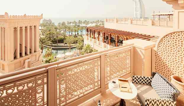 Utsikt från balkong med stolar, bord och arabiska byggnader i förgrunden, samt palmer och Burj Al Arab i bakgrunden. (AI-genererad text)