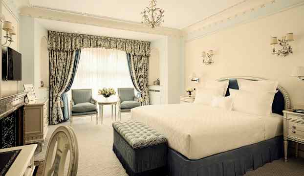 Ett elegant hotellrum med en stor säng, två stolar vid ett bord och rikligt dekorerade gardiner. (AI-genererad text)