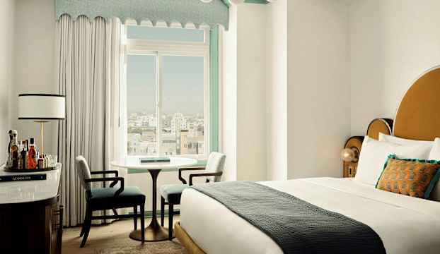 Ett hotellrum med en dubbelsäng, två stolar och ett runt bord vid ett fönster. Det finns också en byrå med drycker och en lampa. (AI-genererad text)