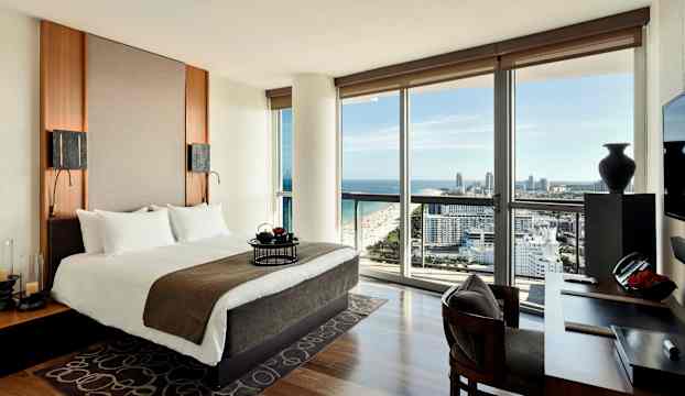 Ett modernt hotellrum med en dubbelsäng, skriftbord och en utsikt över staden och havet genom stora fönster. (AI-genererad text)