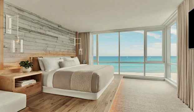 Ett ljust hotellrum med en stor dubbelsäng och utsikt över havet genom stora fönster. (AI-genererad text)