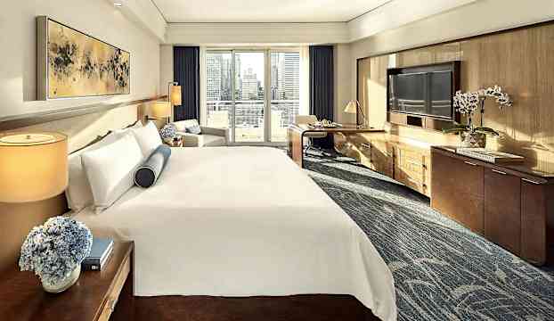 Ett ljus och elegant hotellrum med en stor säng, två sidobord, en soffa och ett skrivbord vid fönstret med utsikt över staden. (AI-genererad text)