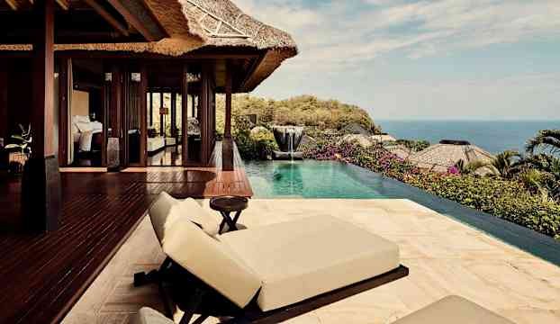 Utsikt över en utomhusterrass med solstolar och pool, omgiven av växtlighet och hav i bakgrunden. (AI-genererad text)