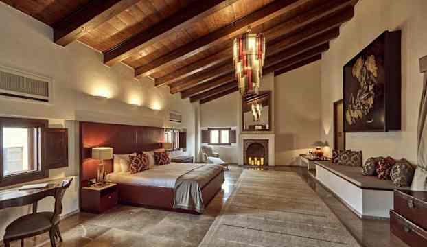 Ett hotellrum med synliga träbjälkar i taket, en stor säng, en öppen spis och eleganta möbler. (AI-genererad text)