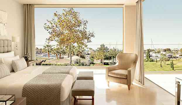 Hotellrum med en stor säng och sittmöbler samt utsikt över en grönskande trädgård och småbåtshamn. (AI-genererad text)