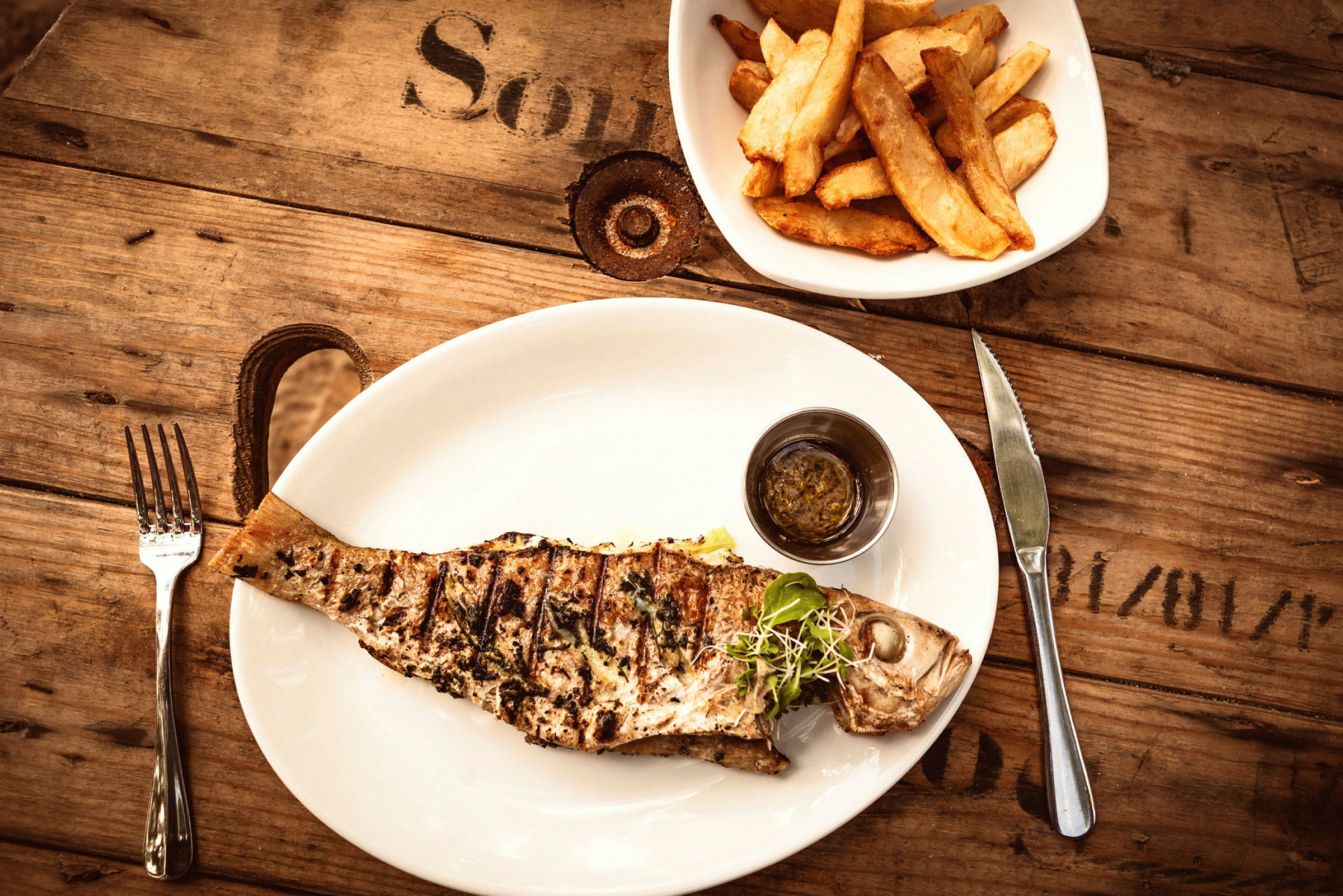 Grillad fisk serverad på en vit tallrik med sås, bredvid en skål med pommes frites på ett träbord. (AI-genererad text)