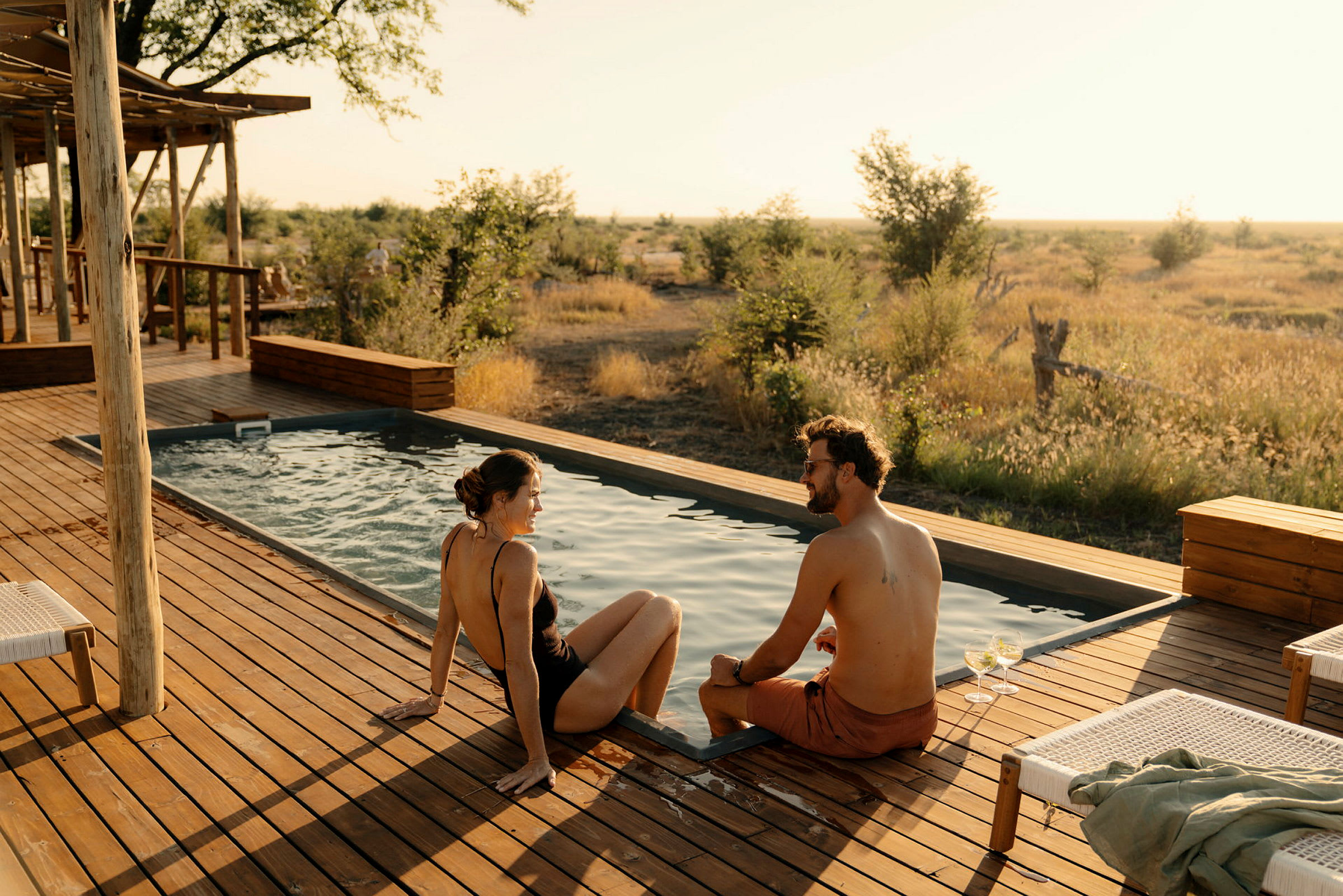 Två personer sitter vid kanten av en pool på en träveranda med utsikt över en gräsbevuxen natur. (AI-genererad text)
