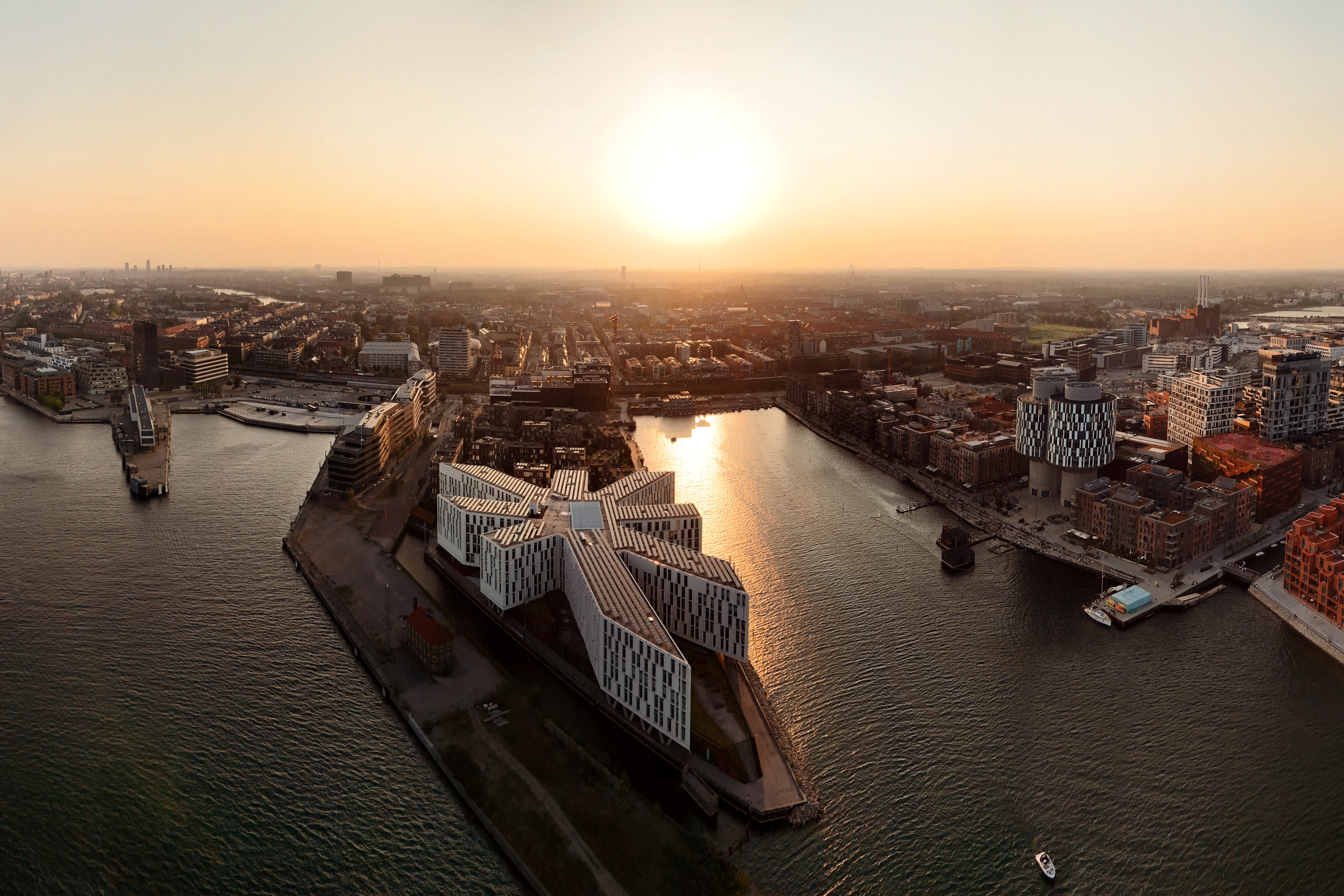 Flygbild över en stad med moderna byggnader vid vattnet och en solnedgång i bakgrunden. (AI-genererad text)