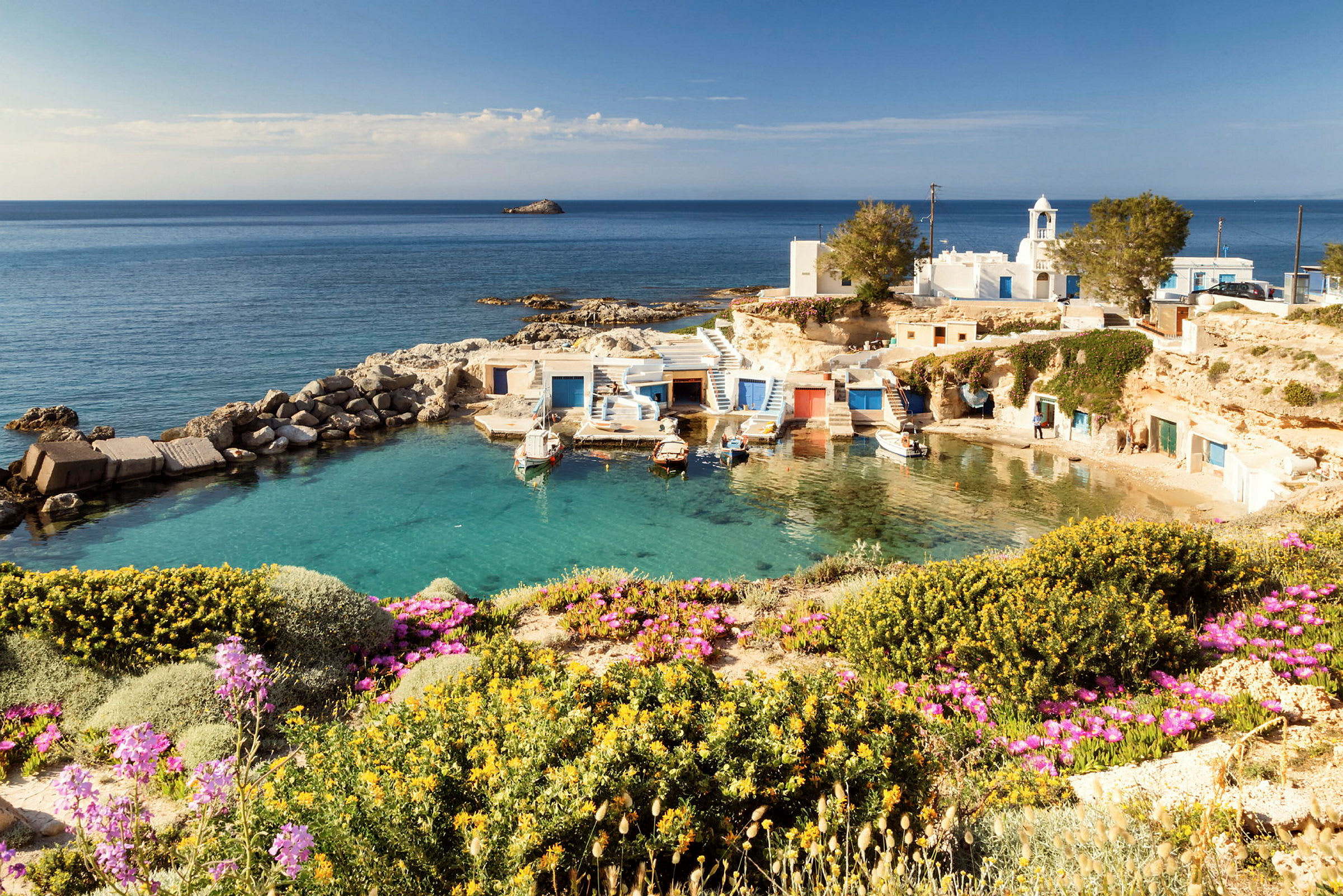 Bild av en liten pittoresk hamn med små båthytter i blå och vita färger vid havet. Förgrunden visar färgglada blommor och grönska. (AI-genererad text)