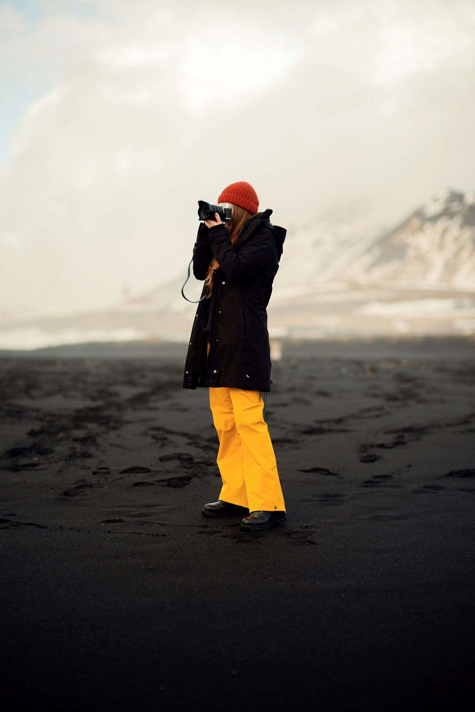 Person i svarta kläder och gul byxdräkt fotograferar på svart sandstrand. (AI-genererad text)