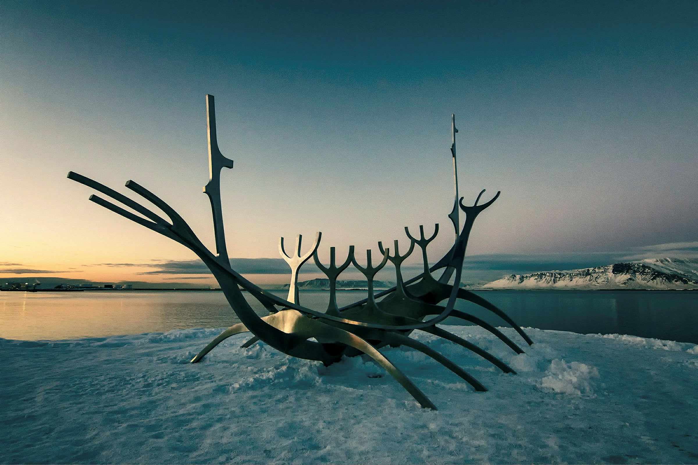 Metallskulpturen av ett stiliserat skepp står på en snötäckt yta vid vattnet. (AI-genererad text)