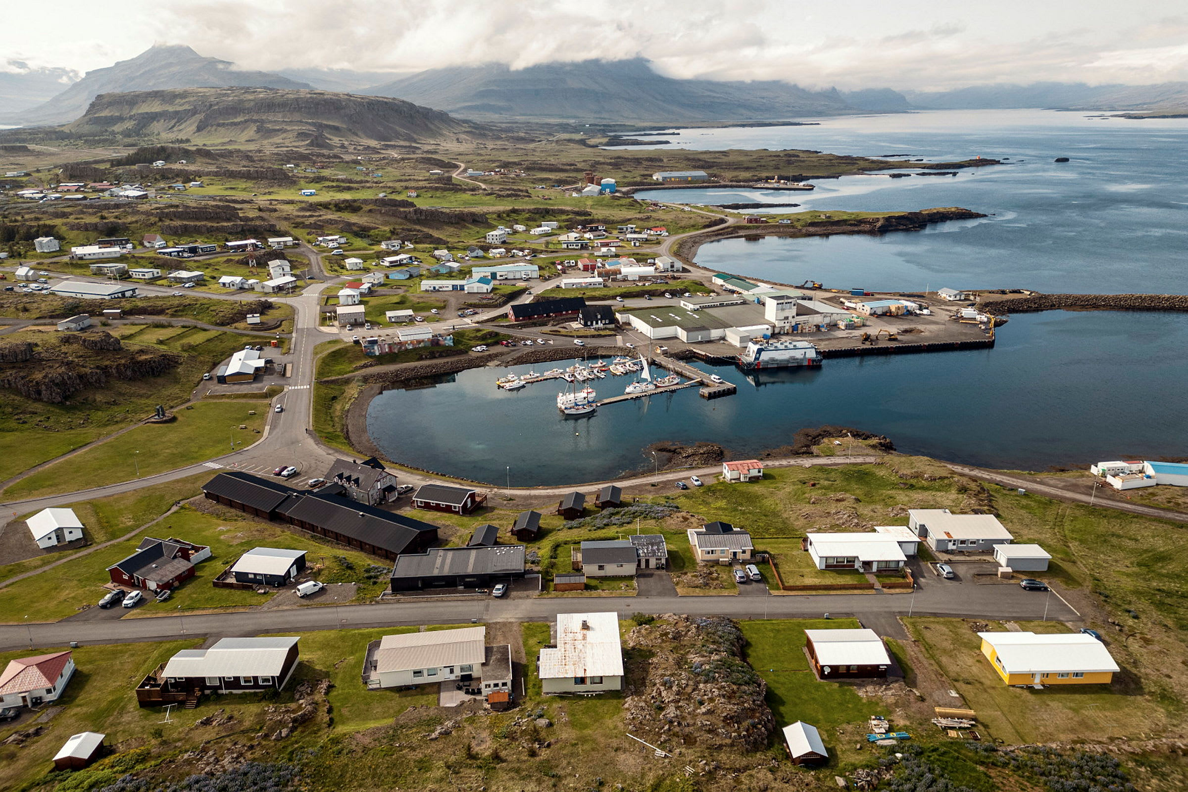 Flygfoto över en liten kuststad med en hamn fylld med båtar, omgiven av gröna fält och hus. (AI-genererad text)