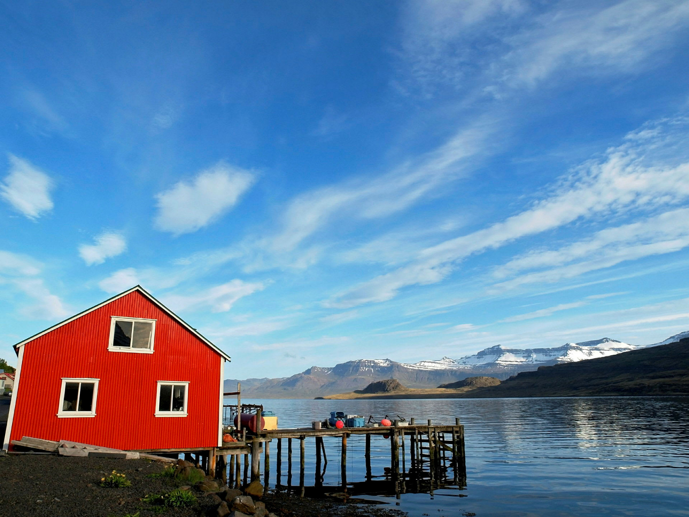 Ett rött hus står vid vattnet bredvid en brygga, med berg och en klarblå himmel i bakgrunden. (AI-genererad text)