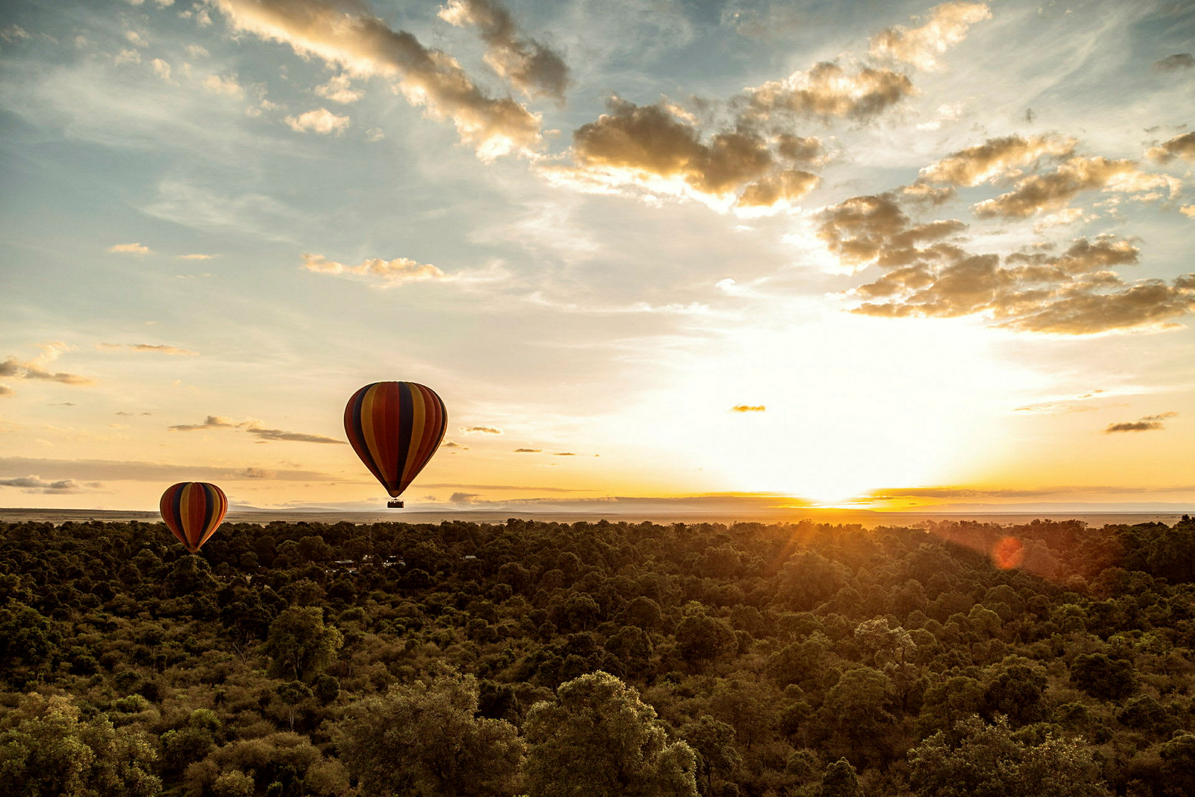 Två färgglada luftballonger flyger över ett skogslandskap vid solnedgång. (AI-genererad text)