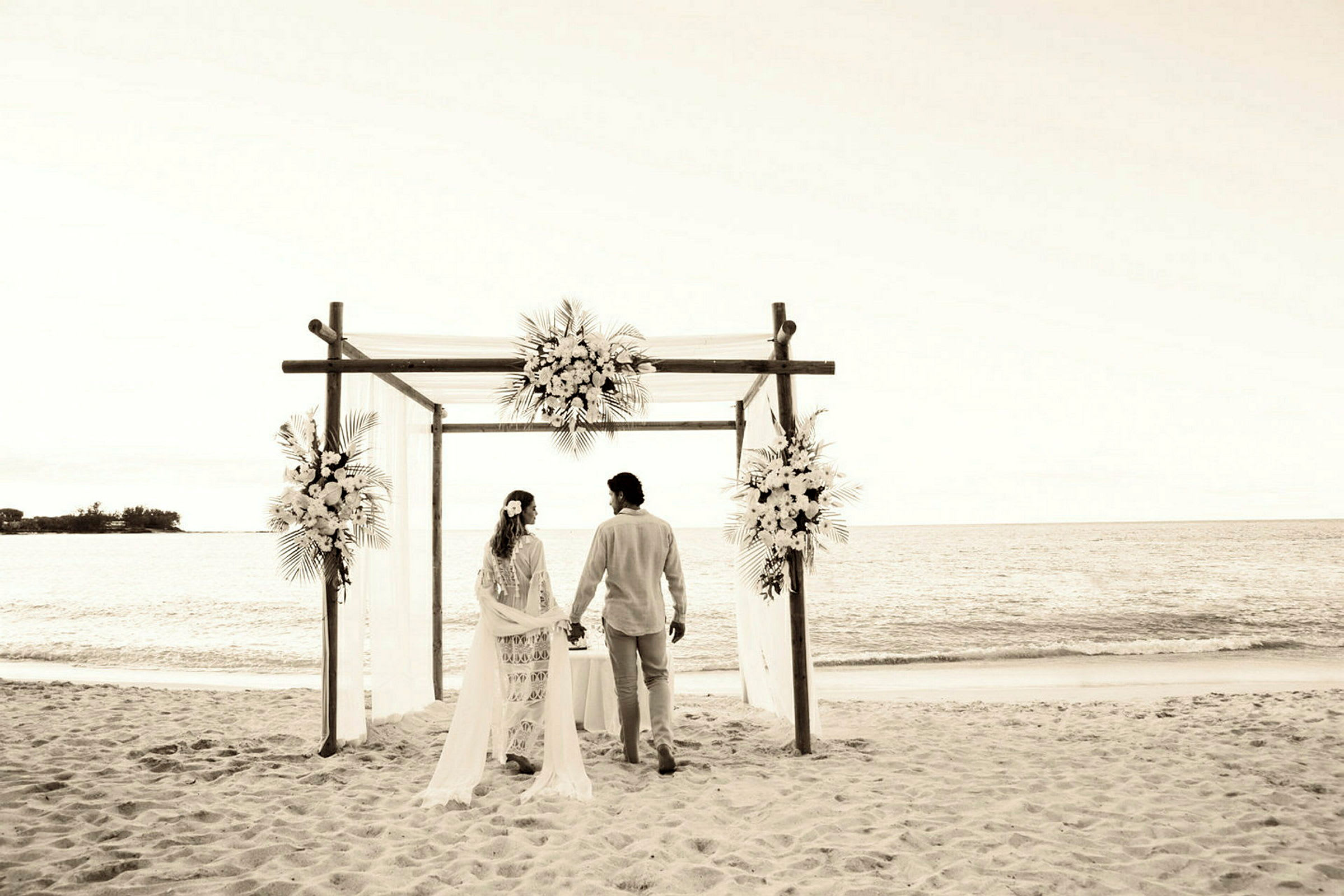 Ett par står under en blomsterprydd portal på en sandstrand med havsutsikt. (AI-genererad text)