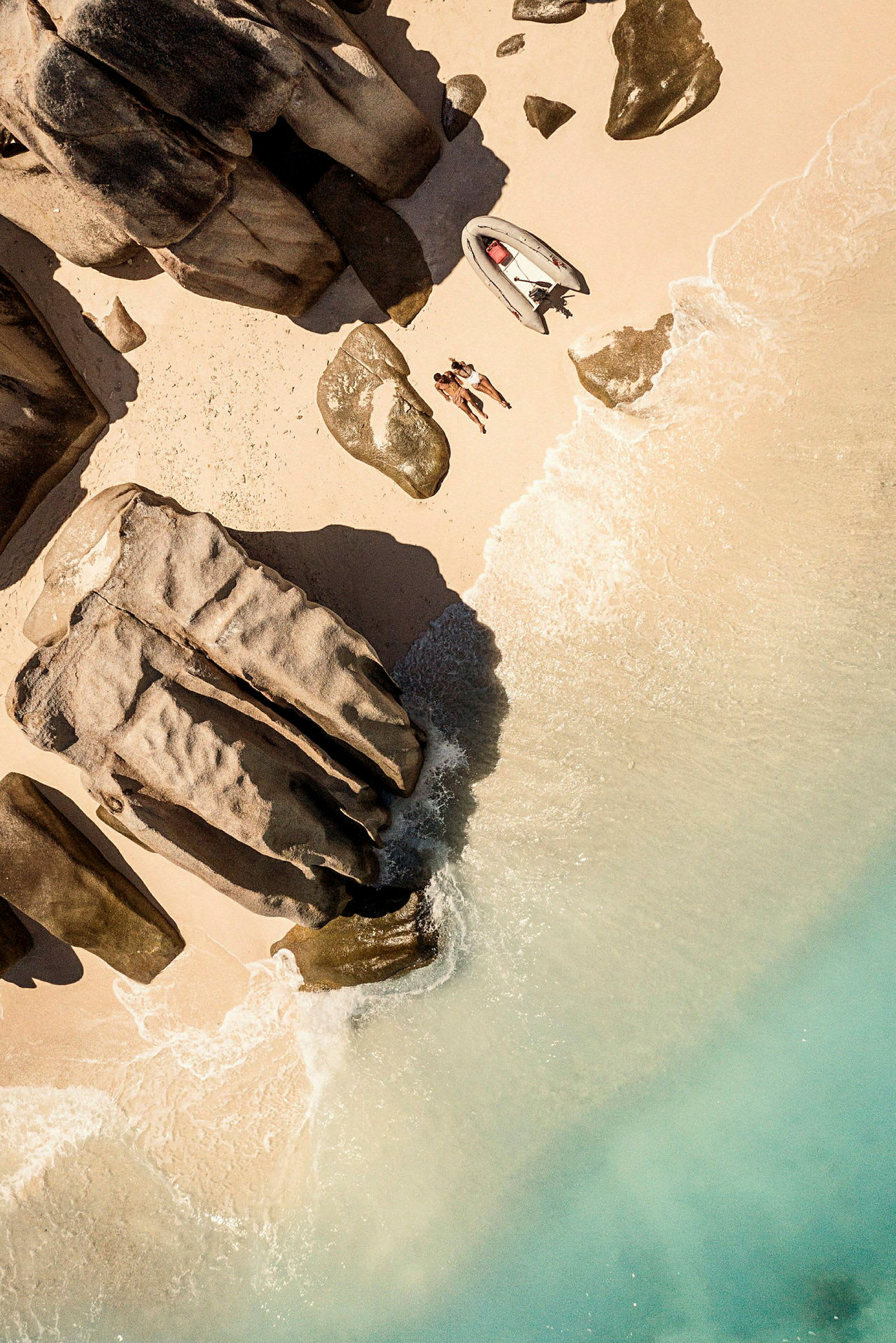 Flygfoto av en sandstrand med stora klippformationer, en båt och två personer som ligger på stranden. (AI-genererad text)