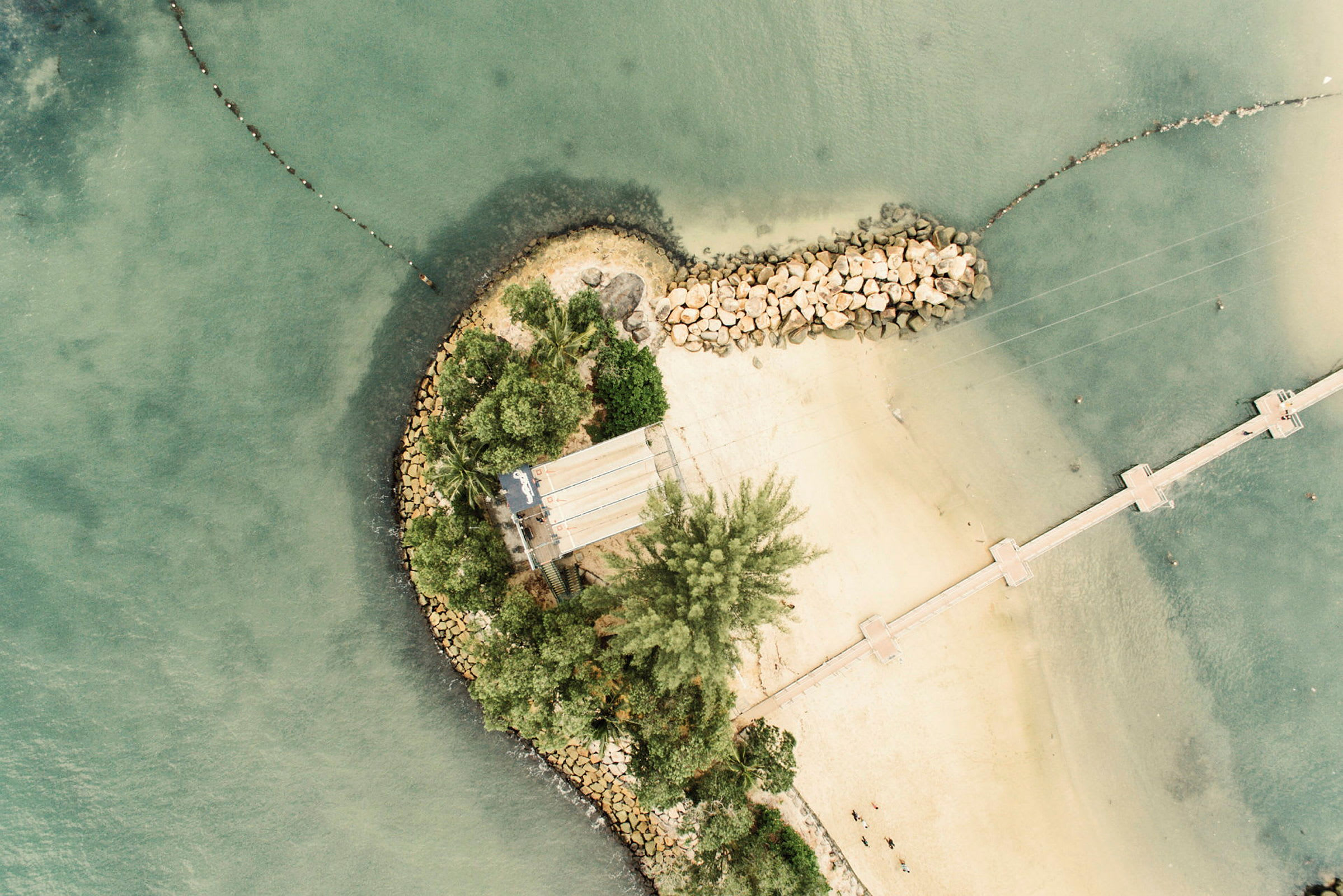 Flygbild av en ö med sandstrand, gröna träd, stenbarriär och brygga som leder till vattnet. (AI-genererad text)