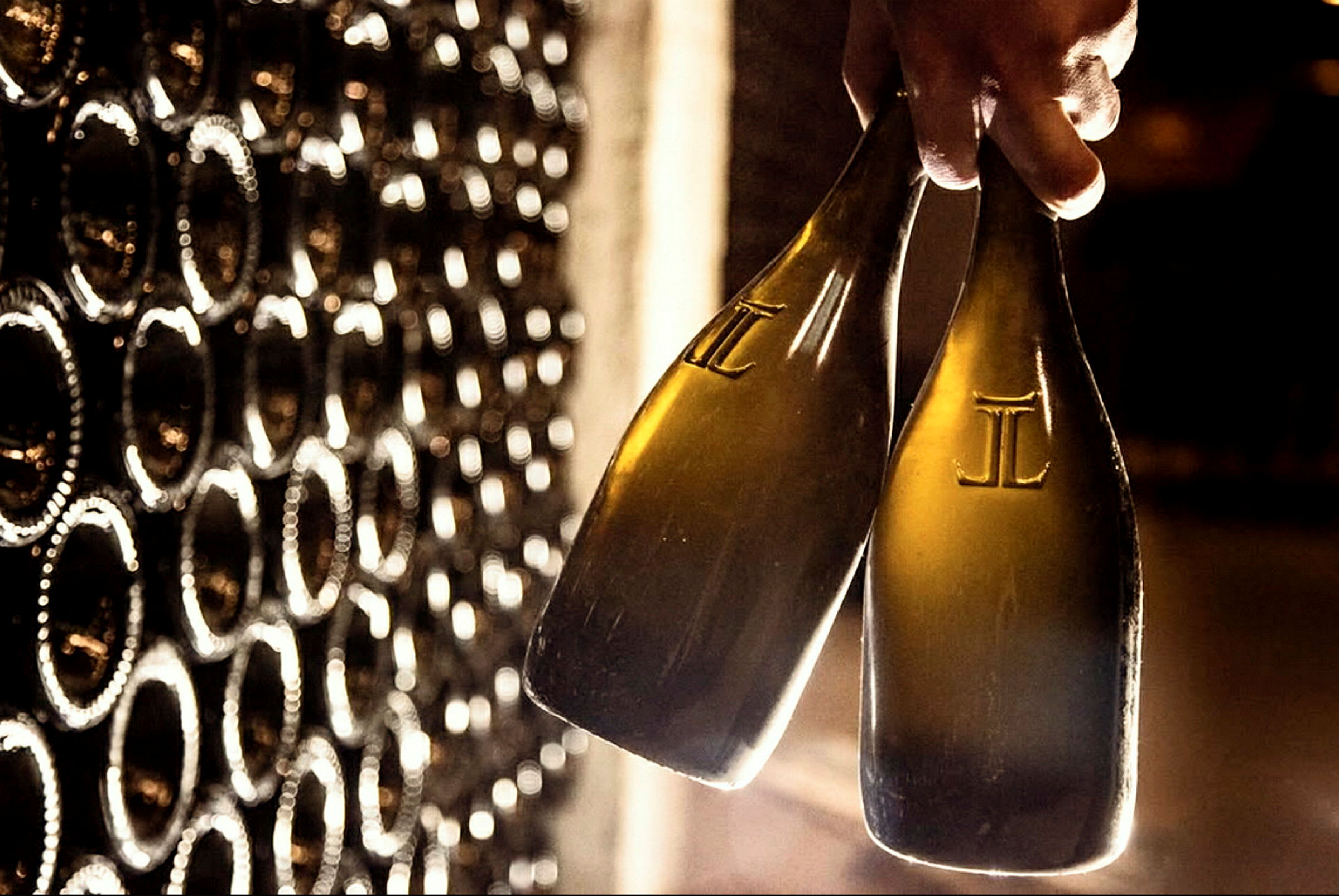 En hand håller två champagneflaskor framför en hylla med flera flaskor. (AI-genererad text)