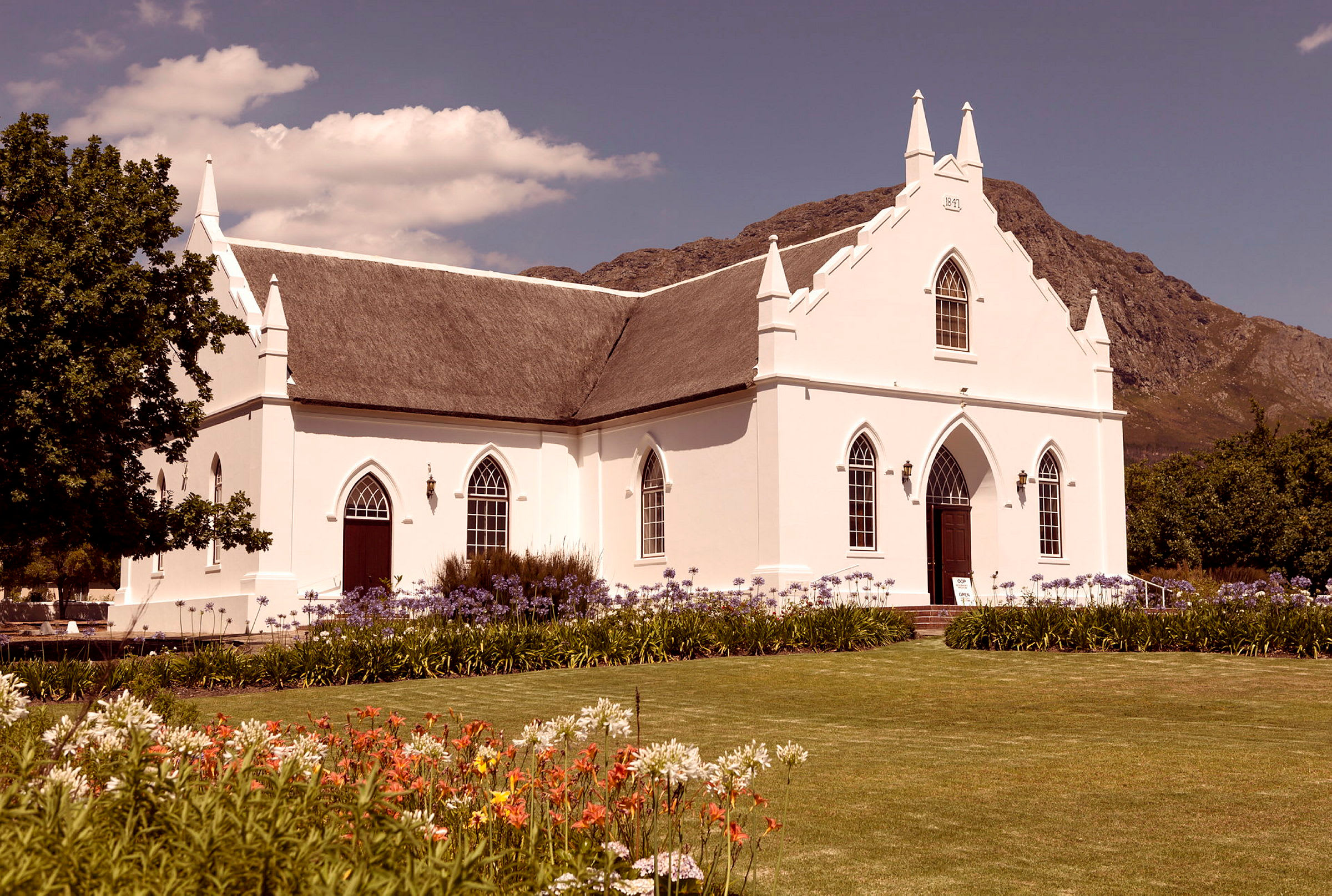 En vit kyrka med spetsiga tak och gotiska fönster, omgiven av blommor och gräsmatta. (AI-genererad text)
