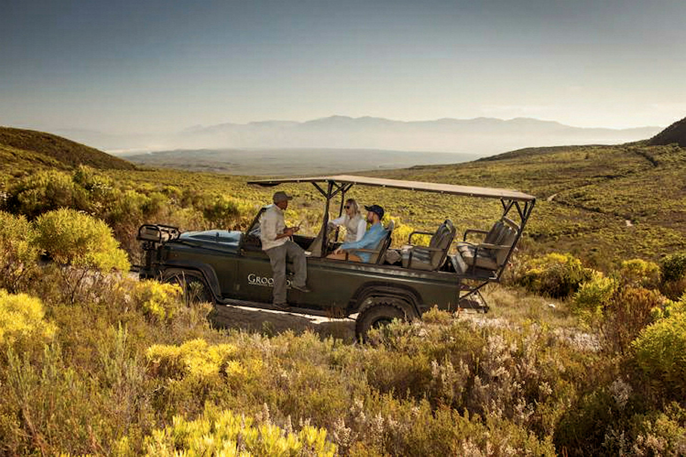 En öppen safari-jeep står bland torra buskar i ett kuperat landskap, med två personer sittande bak och en guide ståendes bredvid. (AI-genererad text)