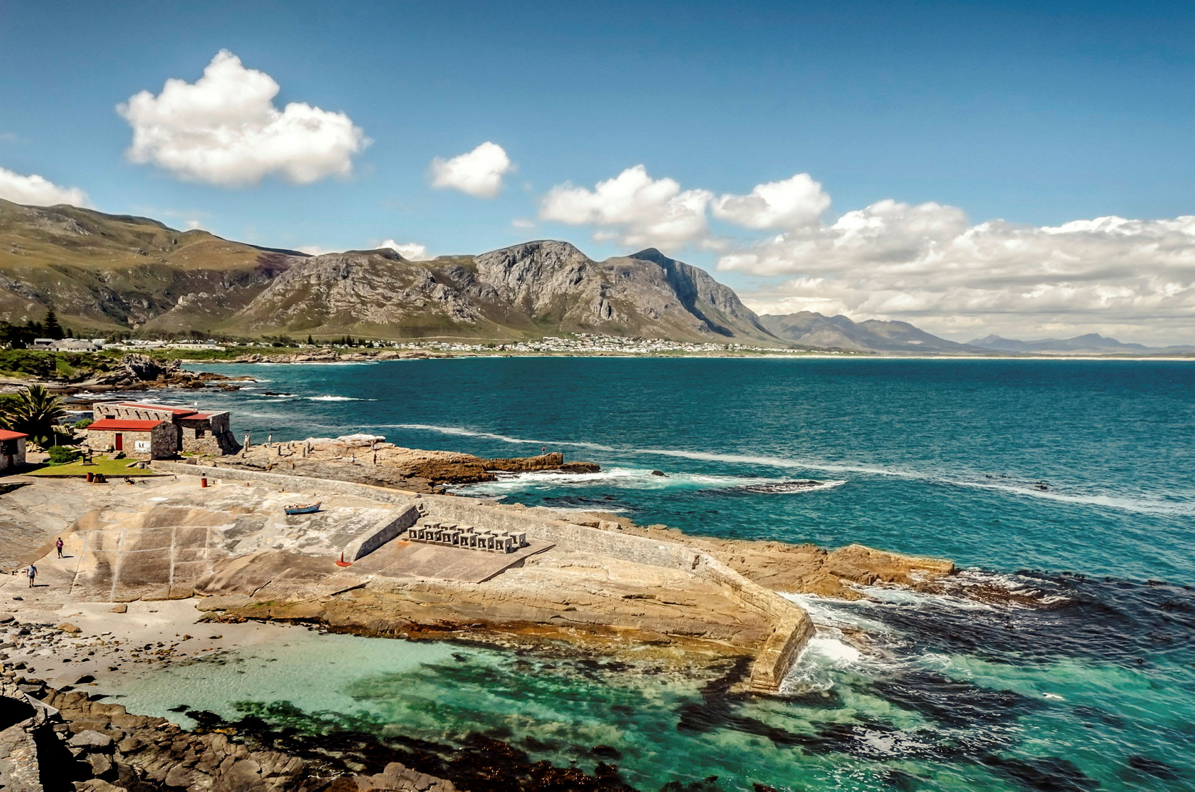 En kustlinje med klart turkost vatten, stenig strand och en byggnad med ett rött tak. Berg syns i bakgrunden under en blå himmel. (AI-genererad text)