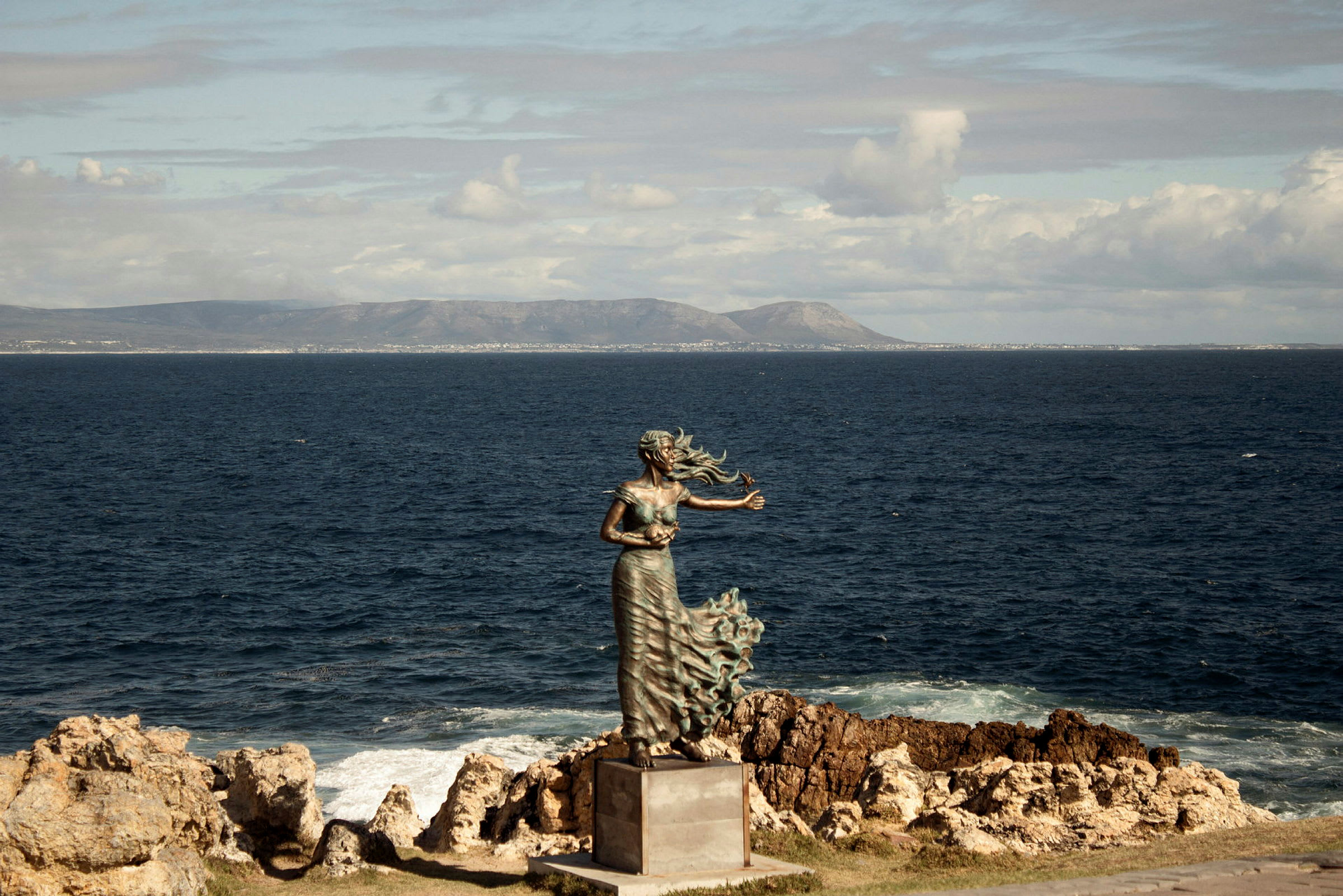 En staty föreställande en kvinna med vågigt hår och draperad klädsel står på en sockel vid havet med klippig kust och berg i bakgrunden. (AI-genererad text)