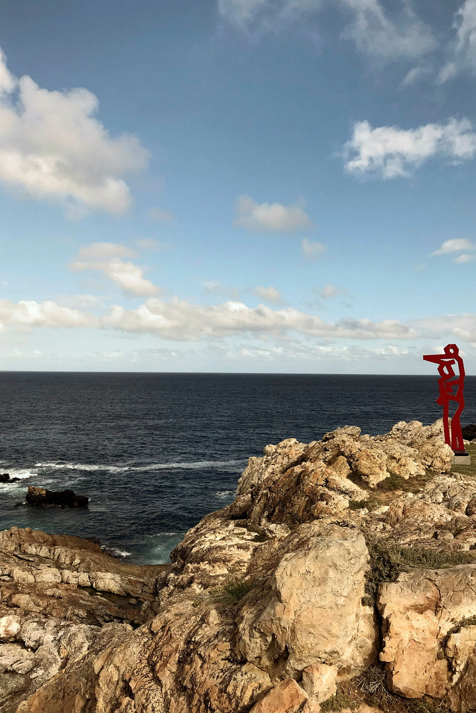 En klippa vid havet under blå himmel där en röd skulptur står på kanten. (AI-genererad text)